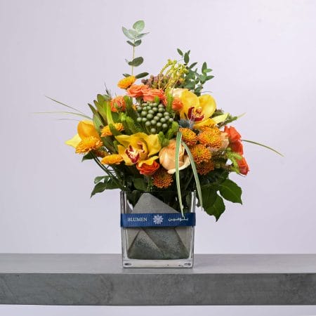 تنسيق زهور أنيق يحتوي على زهور الأوركيد الصفراء والورود البرتقالية وتوت أخضر في مزهرية زجاجية عصرية من بلومن.