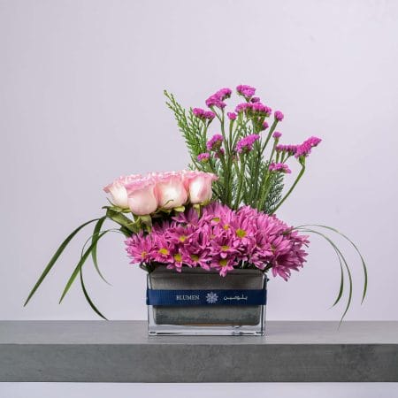 تنسيق زهور راقٍ يضم وروداً وردية وأزهاراً بنفسجية داخل مزهرية زجاجية عصرية من بلومن.