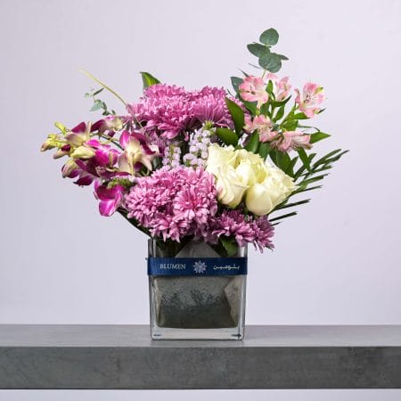 تنسيق زهور أنيق يضم أقحوان بنفسجي ووروداً بيضاء وأزهار ألسْتْرُوميريا وردية في مزهرية زجاجية من بلومن.