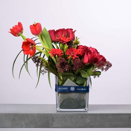 تنسيق زهور أنيق يضم ورودًا حمراء وتوليب وجربيرا في مزهرية زجاجية عصرية من بلومن.