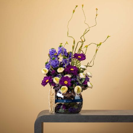 تنسيق زهري بأزهار أرجوانية وبيضاء مع فروع ملتوية طويلة في مزهرية زجاجية مستديرة على خلفية بيج