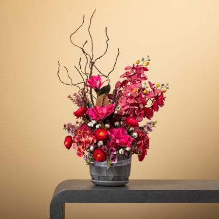 تنسيق زهري بدرجات الوردي والأحمر مع فروع ملتوية طويلة في مزهرية رمادية مزخرفة على خلفية بيج