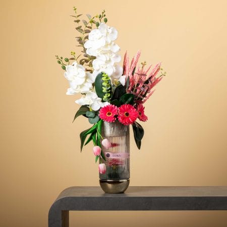 تنسيق زهري فاخر وطويل يضم أوركيد أبيض وجربيرا وردية وتوليب في مزهرية زجاجية مخططة أمام خلفية بيج.