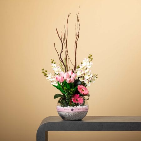 تنسيق زهري طويل وإبداعي يضم أوركيد أبيض وتوليب وردي وجربيرا وردية في مزهرية زجاجية دائرية مليئة بالحصى من تصميم بلومن أمام خلفية بيج.