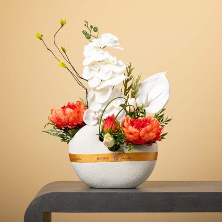 مزهرية دائرية بيضاء ذات سطح خشن تحتوي على أوركيد أبيض متدلٍ وبيوني برتقالي زاهٍ وزهرة أنثوريوم بيضاء مع خضار طبيعي، بتنسيق من بلومن أمام خلفية بيج.