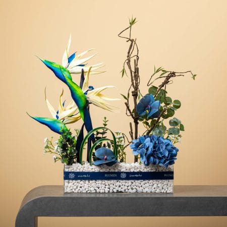 مزهرية زجاجية مستطيلة مليئة بالحصى البيضاء تحتوي على زهور تشبه طائر الفردوس بألوان الأزرق والأخضر مع أوركيد أزرق وهدرانجيا زرقاء وأغصان ملتوية وخضار طبيعي، بتنسيق من بلومن أمام خلفية بيج.