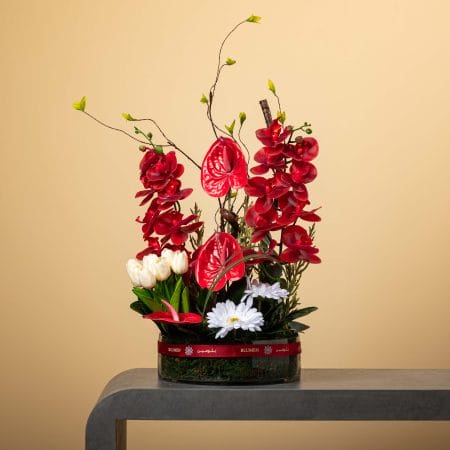 مزهرية زجاجية بيضاوية مزينة بشريط أحمر، تحتوي على أوركيد أحمر وزهور أنثوريوم حمراء وتوليب أبيض وأقحوان أبيض مع خضار طبيعي وأغصان فنية، بتنسيق من بلومن أمام خلفية بيج.