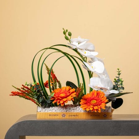 مزهرية مستطيلة مليئة بالحصى البيضاء تضم أقواساً خضراء من الأوراق مع أوركيد أبيض متدلٍ وجربيرا برتقالية زاهية وخضار طبيعي، مزينة بشريط أصفر من بلومن أمام خلفية بيج.