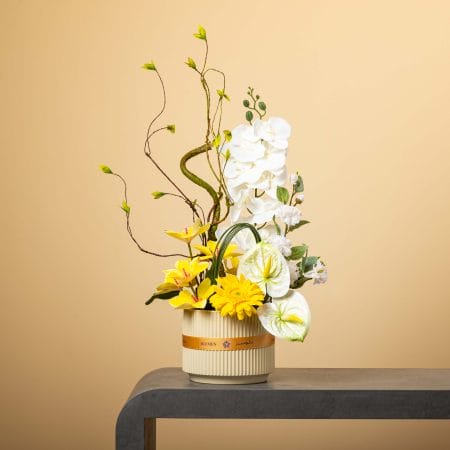 وعاء كريمي مخطط مزين بشريط برتقالي من بلومن، يحتوي على أوركيد أبيض متدلٍ وزنابق وجرابيرا صفراء وأنثوريوم أبيض مائل للأخضر مع أغصان ملتوية وخضار طبيعي أمام خلفية بيج.