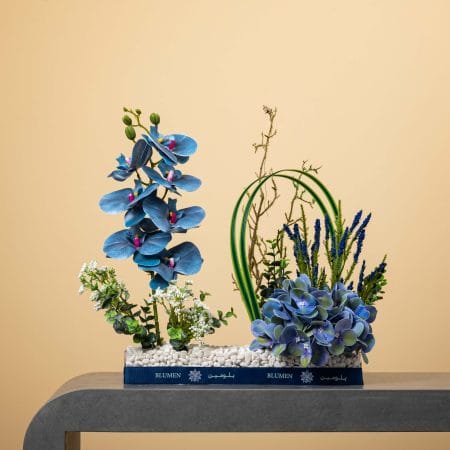 مزهرية مستطيلة مليئة بالحصى البيضاء ومزينة بشريط كحلي من بلومن، تحتوي على أوركيد أزرق وهدرانجيا زرقاء وأوراق منحنية خضراء وزهور زرقاء رفيعة مع أغصان وخضار طبيعي أمام خلفية بيج.