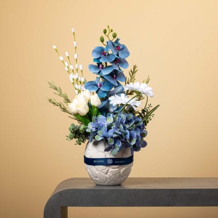 تنسيق زهري بأزهار زرقاء وبيضاء يضم أوركيد وهدرانج وتوليب في مزهرية بيضاء مستديرة ذات نقش بارز مع شريط كحلي على خلفية بيج