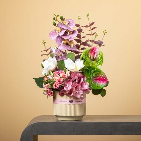 تنسيق زهري ناعم بأزهار وردية وبيضاء مع أوركيد، هدرانج، ورود وأنثوريوم أخضر في مزهرية بيج مخططة على خلفية بيج