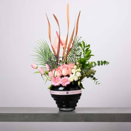 مزهرية سوداء دائرية مخططة مزينة بشريط وردي من بلومن، تحتوي على ورود وردية ناعمة وجربيرا وردية وتوليب وردي وزهور بيضاء مع أوراق نخيل وخضار طبيعي وسيقان بنية ديكورية طويلة أمام خلفية رمادية فاتحة.