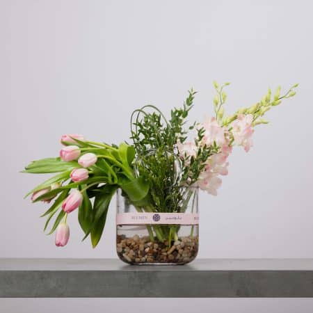 مزهرية زجاجية شفافة على شكل جرة تحتوي على ماء وحصى نهرية، منسّقة بتوليب وردي متدلٍ وزهور وردية ناعمة مع خضار طبيعي، ومزينة بشريط وردي من بلومن أمام خلفية رمادية فاتحة.