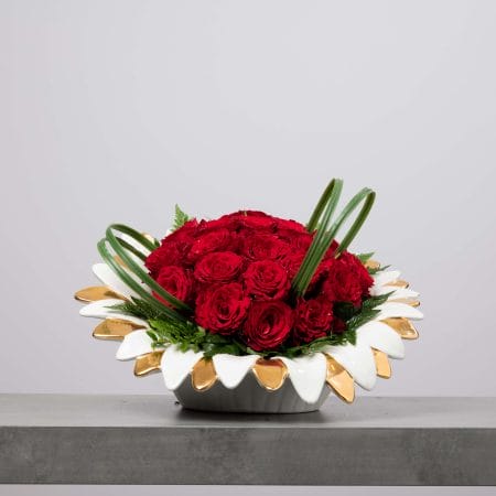 وعاء خزفي منخفض على شكل بتلات بيضاء بحواف ذهبية، مملوء بقبة كثيفة من الورود الحمراء مع أوراق خضراء وأوراق عشب منحنية، موضوع على سطح رمادي أمام خلفية رمادية فاتحة.