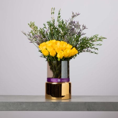 مزهرية زجاجية طويلة بقاعدة ذهبية مزينة بشريط بنفسجي من بلومن، مليئة بورود صفراء زاهية وخضار كثيف وزهور بنفسجية ناعمة أمام خلفية رمادية فاتحة.