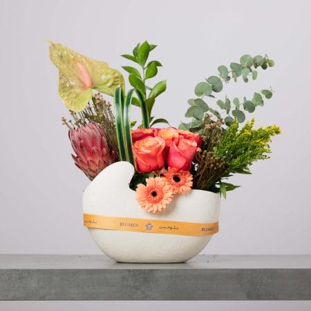 مزهرية بيضاء مجسّمة على شكل وعاء مزينة بشريط أصفر من بلومن، منسقة بورود برتقالية مائلة للزهري وجربيرا خوخية وأنثوريوم أخضر وبروتيا وردية وأوكالبتوس وخضار متنوع أمام خلفية رمادية فاتحة.