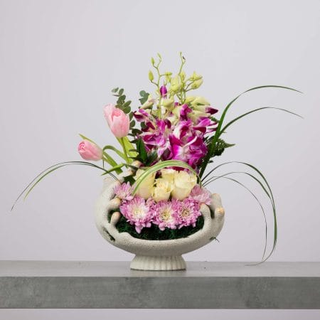 مزهرية وعائية مرتفعة بلون أبيض مائل للعاج وبملمس مجسّم، منسّقة بتوليب وردي وأوركيد بنفسجي وأورود بيضاء وأقحوان وردي فاتح مع أوراق عشبية طويلة وأوكالبتوس وخضار طبيعي أمام خلفية رمادية فاتحة.