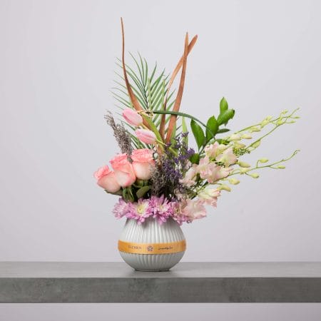 مزهرية دائرية مخططة باللون الأبيض مزينة بشريط ذهبي من بلومن، منسقة بورود وردية ناعمة وتوليب وردي وأوركيد فاتح وزهور بنفسجية صغيرة وأقحوان مع ورقة نخيل وخضار طبيعي أمام خلفية رمادية فاتحة.