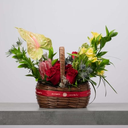 سلة خوص بنية مزينة بشريط أحمر من بلومن، مملوءة بورود حمراء وزهور صفراء وأنثوريوم أخضر وبروتيا وردية وشوك أزرق وخضار كثيف أمام خلفية رمادية فاتحة.