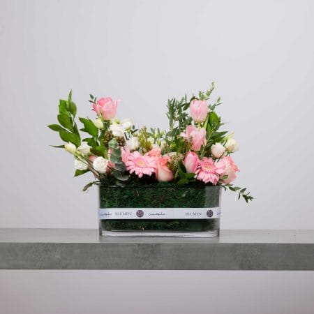 Low rectangular glass vase wrapped with a white Blumen ribbon, filled with pink tulips, pink gerbera daisies, blush roses, white blooms, greenery, and moss against a light grey background.