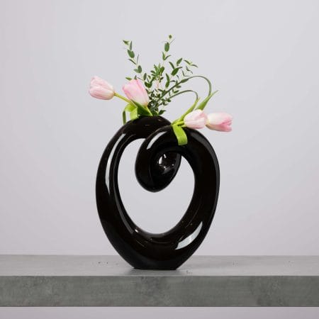 مزهرية سوداء مجسّمة على شكل قلب بحركة لولبية عصرية، بداخلها توليب وردي ناعم مع خضار خفيف، موضوعة على سطح رمادي أمام خلفية رمادية فاتحة.