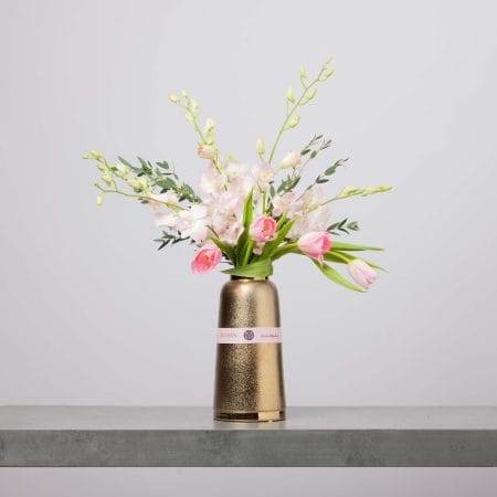 مزهرية طويلة بلون ذهبي معدني مزينة بشريط وردي من بلومن، تحتوي على توليب وردي ناعم وأوركيد وردي فاتح وبراعم خضراء وخضار خفيف، أمام خلفية رمادية فاتحة.