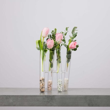 أربع مزهريات أنبوبية طويلة شفافة متصلة ببعضها، يحتوي كل منها على حصى ملوّن في القاعدة مع توليب وردي ناعم وأغصان أوكالبتوس خضراء، موضوعة على سطح رمادي أمام خلفية رمادية فاتحة.
