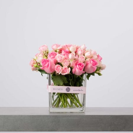 مزهرية زجاجية شفافة مربعة مزينة بشريط وردي من بلومن، مليئة بطبقات من الورود والروزات الصغيرة باللون الوردي على شكل قبة كثيفة من الأزهار أمام خلفية رمادية فاتحة.