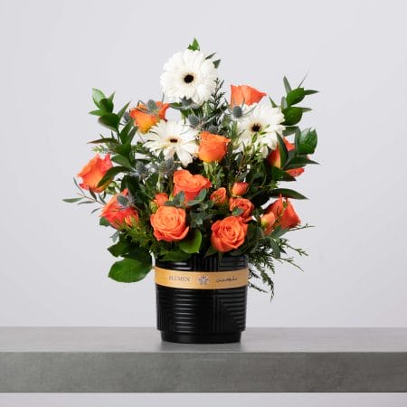 مزهرية أسطوانية سوداء مخططة مزينة بشريط ذهبي من بلومن، منسقة بورود برتقالية زاهية وجربيرا بيضاء مع لمسات من الشوك الأزرق وخضار كثيف أمام خلفية رمادية فاتحة.