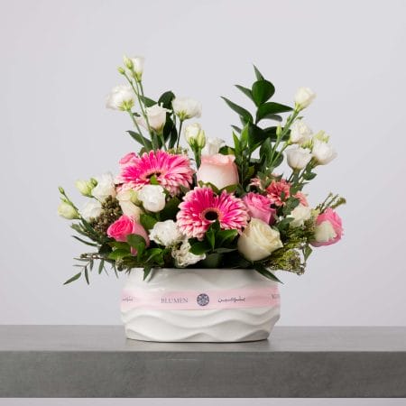 مزهرية خزفية بيضاء منخفضة ذات تموّجات ناعمة مزينة بشريط وردي من بلومن، مليئة بجربيرا وردية زاهية وورود وردية وبيضاء وليزيانثس أبيض مع خضار كثيف أمام خلفية رمادية فاتحة.