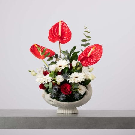 مزهرية على شكل وعاء مرتفع بتفاصيل بيضاء خشنة، منسقة بثلاثة أزهار أنتوريوم حمراء طويلة، وجربيرا بيضاء، وورود حمراء، وزهور بيضاء، وشوكيات زرقاء وخضار كثيف أمام خلفية رمادية فاتحة.