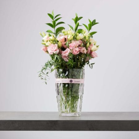 مزهرية زجاجية طويلة مموجة مليئة بزهور الليزيانثس الوردية والكريمية مع أوراق خضراء طبيعية بتنسيق ناعم وانسيابي أمام خلفية رمادية فاتحة.