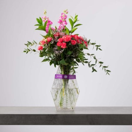 مزهرية زجاجية طويلة بتصميم هندسي يحيط بها شريط بنفسجي، مليئة بورود سبراي وردية زاهية مع أنفاس التنفس الفوشيا وزهور بنفسجية ناعمة وخضار منسدل، بتنسيق طبيعي وانسيابي أمام خلفية رمادية فاتحة.