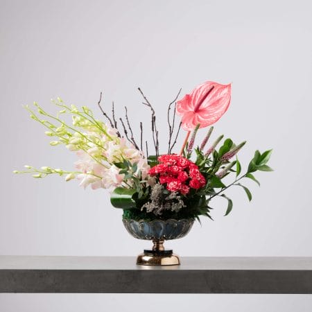 تنسيق زهري غير متماثل في وعاء زجاجي أزرق على قاعدة ذهبية، يضم عناقيد من الأوركيد الباهت المتدلّي من الجهة اليسرى مع زهرة أنثوريوم كورالية واحدة وورود سبراي وردية زاهية وفروع جافة وخضار طبيعي أمام خلفية رمادية فاتحة.