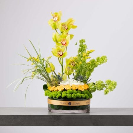 مزهرية زجاجية دائرية تحيط بها طبقة منخفضة من أقحوان أخضر وزهور وردية صفراء، تعلوها زهور بيضاء وسيمبيديوم أوركيد ليموني طويل مع بيلز أوف آيرلند وخضار ناعم بتدرجات أصفر وأخضر مشرقة أمام خلفية رمادية فاتحة.