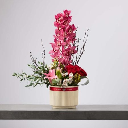 مزهرية كريمية مخططة تحتوي على عمود مرتفع من أوركيد سيمبيديوم وردي مع باقة من الورود الحمراء والزنابق الوردية والزهور البيضاء وأغصان جافة وخضار كثيف بتنسيق عمودي معماري أمام خلفية رمادية فاتحة.