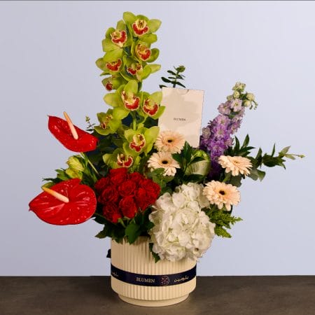 Luxury floral arrangement featuring green orchids, red anthuriums, red roses, white hydrangea, and pastel blooms in an elegant round container, styled for Ramadan.