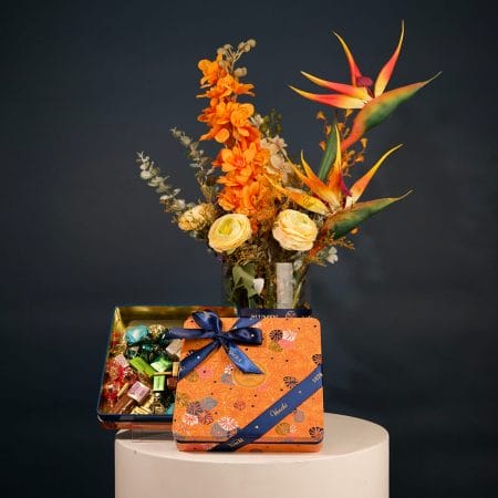 Tropical floral arrangement featuring orange orchids, bird of paradise flowers, and cream roses with greenery in an elegant vase.