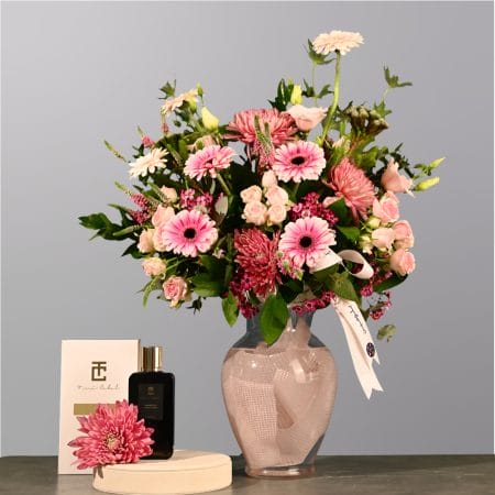 Pink floral arrangement with gerberas, spray roses, and greenery in a soft pink glass vase wrapped with fabric.