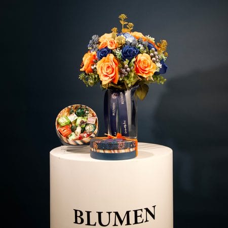 Floral arrangement with bright amber-orange roses, deep blue flowers, and greenery arranged in a clear glass vase with decorative ribbon.