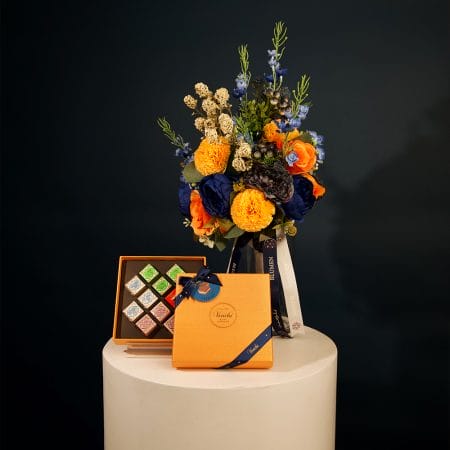 Colorful floral arrangement with golden yellow pom-pom flowers, orange roses, and deep blue blooms arranged in a clear glass vase with decorative ribbons.