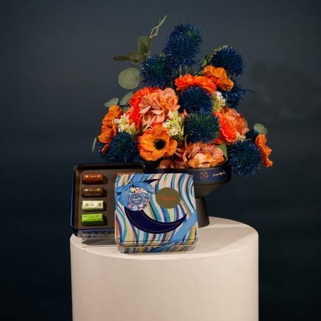 Elegant floral arrangement featuring deep blue spherical flowers and coral-toned blossoms in a modern black pedestal vase.