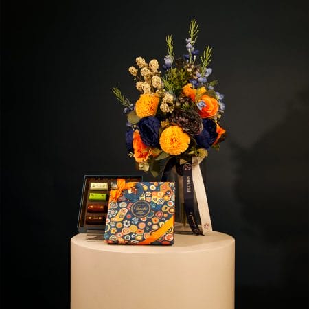 Colorful floral arrangement with golden yellow pom-pom flowers, orange roses, and deep blue blooms arranged in a clear glass vase with decorative ribbons.