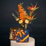 Tropical floral arrangement featuring orange orchids, bird of paradise flowers, and cream roses with greenery in an elegant vase.