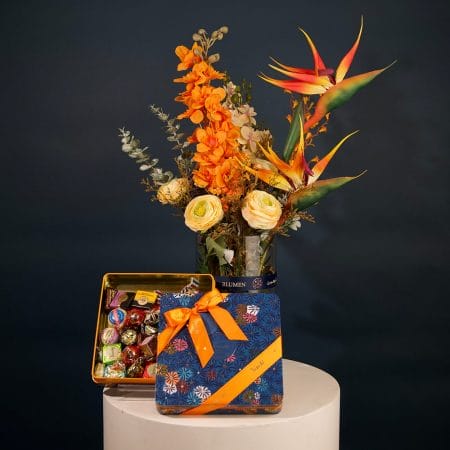 Tropical floral arrangement featuring orange orchids, bird of paradise flowers, and cream roses with greenery in an elegant vase.