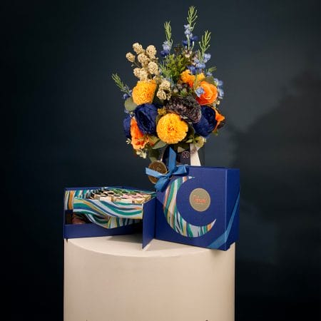 Colorful floral arrangement with golden yellow pom-pom flowers, orange roses, and deep blue blooms arranged in a clear glass vase with decorative ribbons.