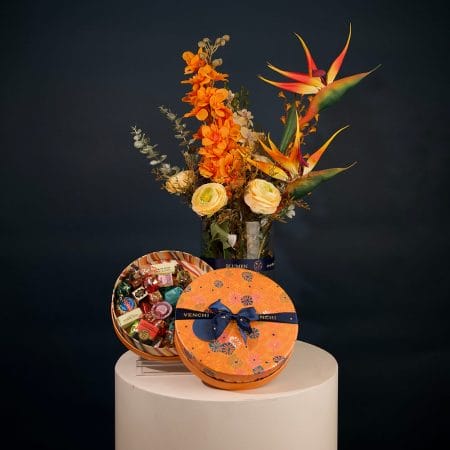 Tropical floral arrangement featuring orange orchids, bird of paradise flowers, and cream roses with greenery in an elegant vase.