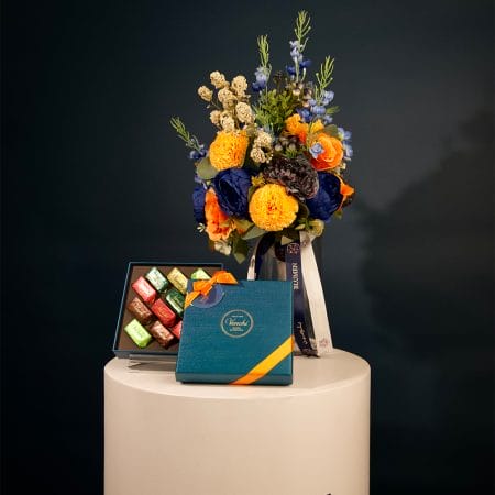 Colorful floral arrangement with golden yellow pom-pom flowers, orange roses, and deep blue blooms arranged in a clear glass vase with decorative ribbons.