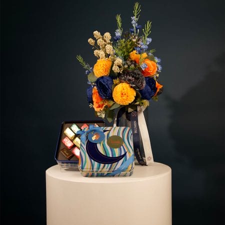 Colorful floral arrangement with golden yellow pom-pom flowers, orange roses, and deep blue blooms arranged in a clear glass vase with decorative ribbons.